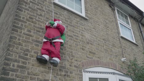 a funny depiction of santa claus toy climbing a rope that hanging from a brick house window during christmas time.