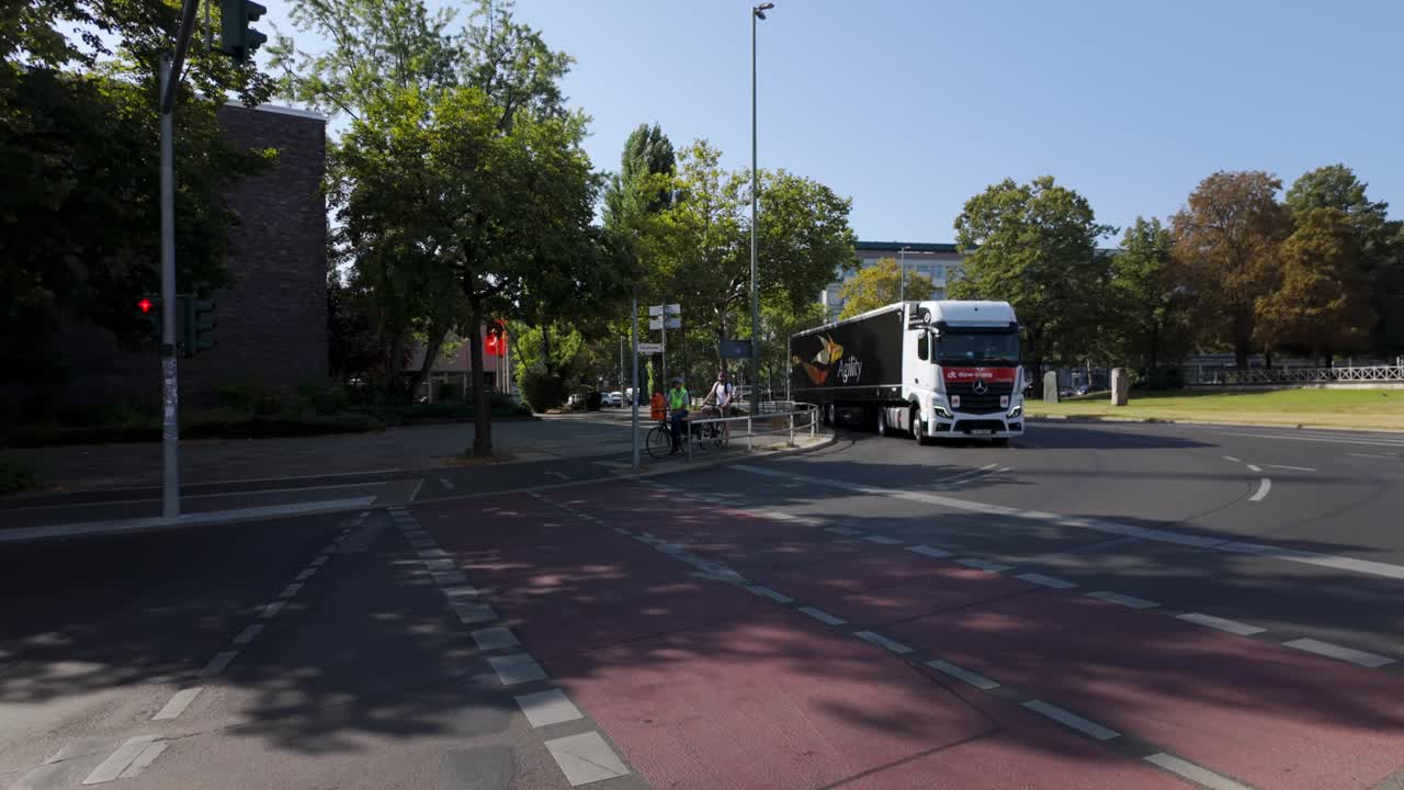 Wide street view in Berlin with a large Agility logistics truck driving past and a man standing on the sidewalk, engrossed in his phone. The camera pans left, capturing city life and traffic