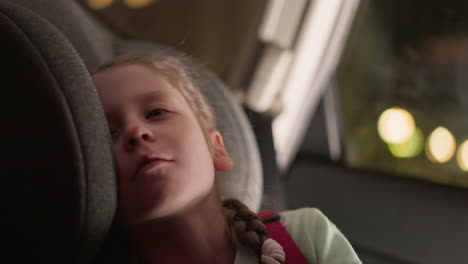 a close view of a child resting her head on the car seat, looking content and relaxed, the blurred lights through the car window