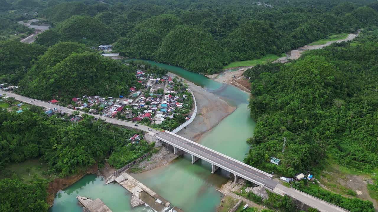 Bridge crossing over green river with vehicles moving along in Virac, surrounded by trees with homes along riverbank
