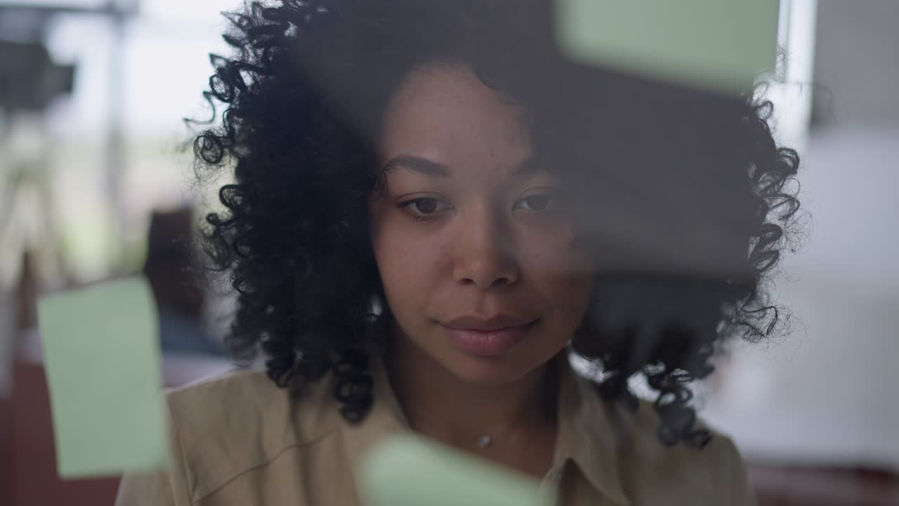 Headshot portrait of gorgeous confident African American woman standing behind glass door analyzing ideas on sticky notes. Close-up face of thoughtful beautiful startuper planning in home office.