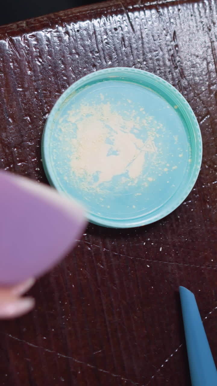 Tapa turquesa con polvos sueltos, mano presionando una esponja morada contra los polvos sobre una mesa de madera, primer plano cenital que muestra el polvo y la textura fina, movimiento suave de golpeteo, escena de preparación de belleza con maquillaje