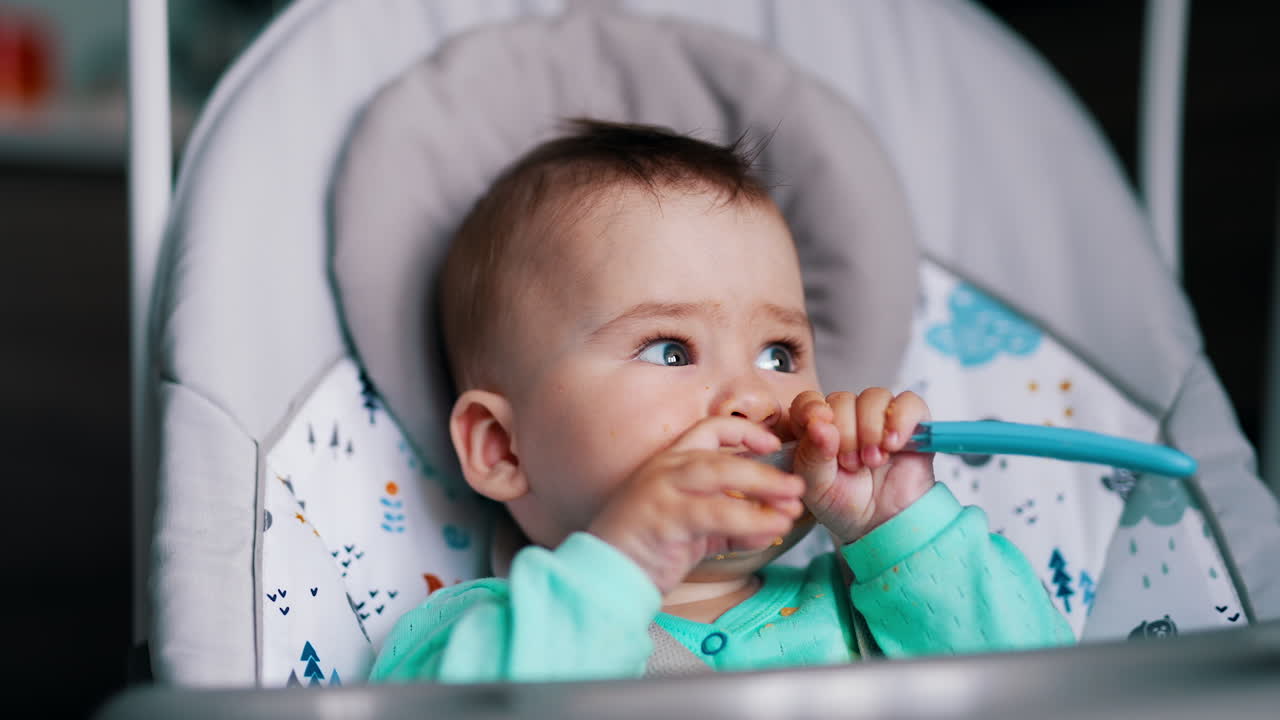 Lovely little boy sitting in a feeding chair. Cute child holds a spoon in his hands and pulls it to his mouth. Close up.