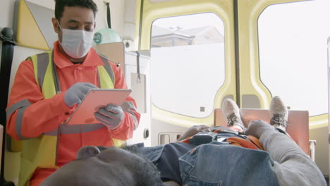 Männlicher-Sanitäter-Mit-Gesichtsmaske-Mit-Tablet-Computer-Während-Der-Fahrt-In-Einem-Krankenwagen