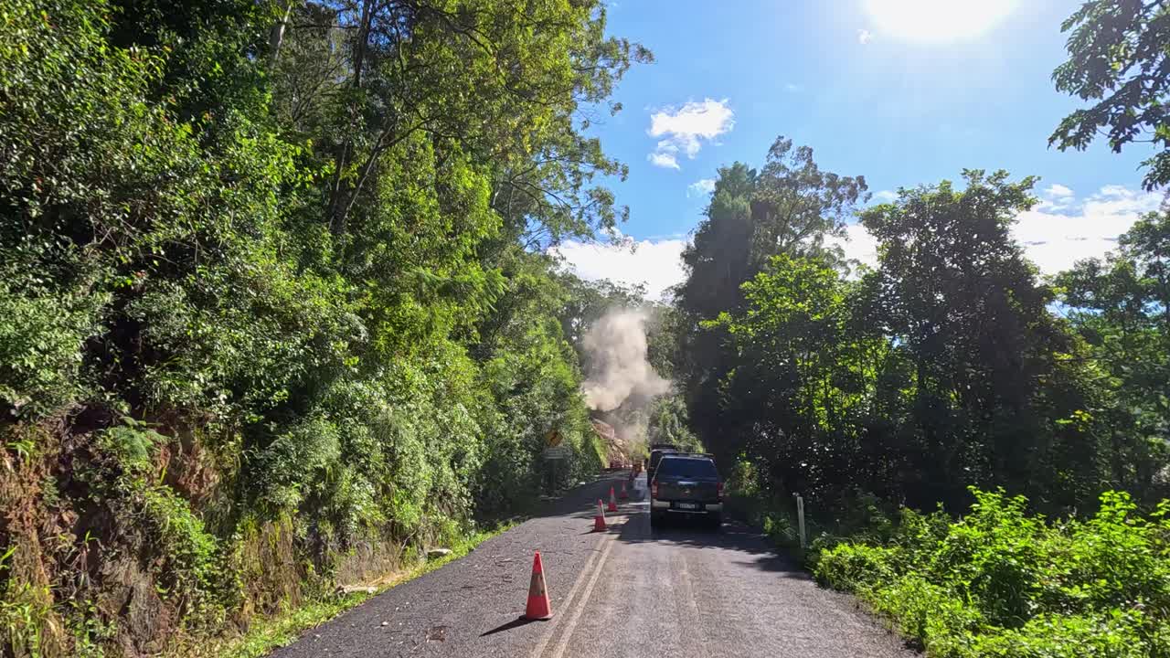A car travels through a lush, sunlit mountain road with dust clouds, surrounded by dense greenery and traffic cones