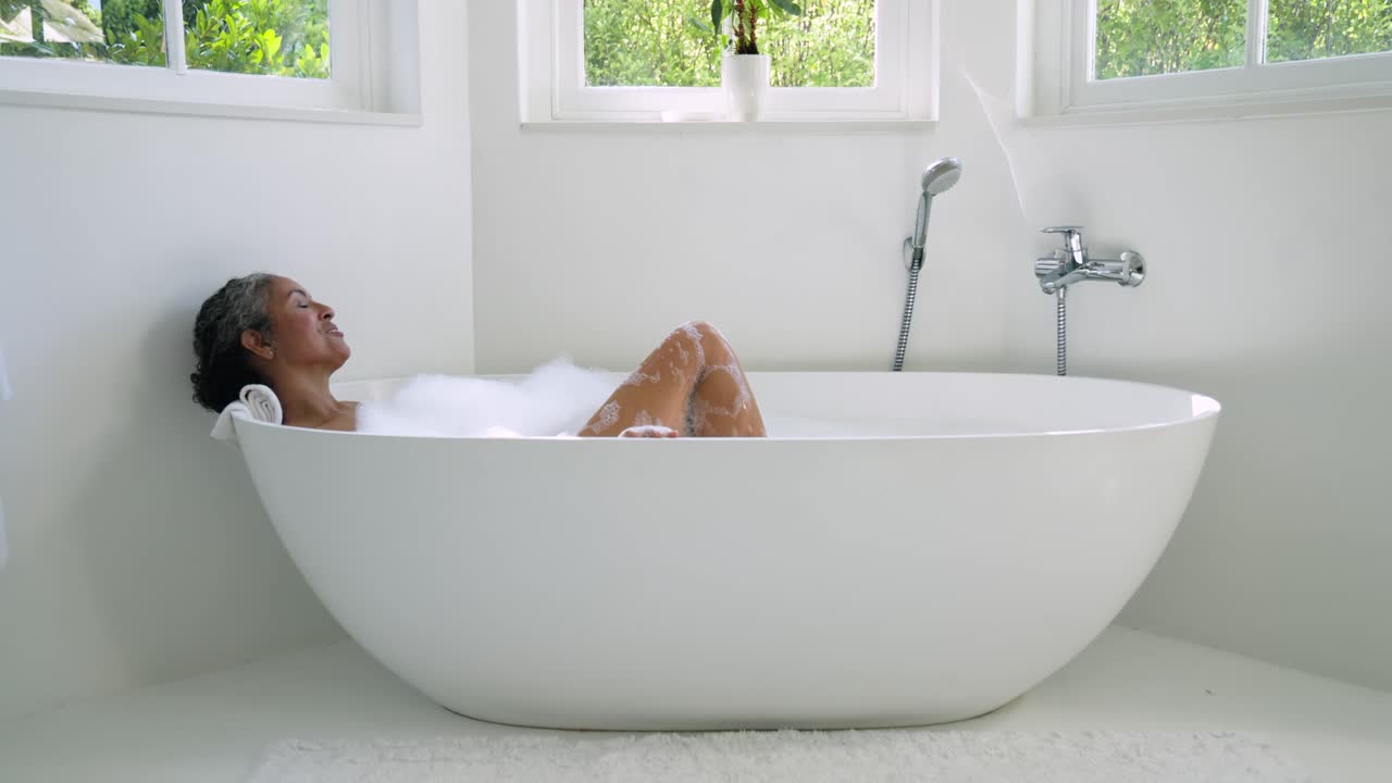 Donna africana anziana che si rilassa nella vasca da bagno vicino alle finestre, muovendo le ginocchia mentre l'acqua scorre dal rubinetto e le bolle si formano.