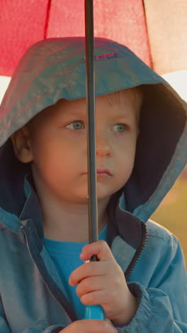 upset kid with umbrella at rainfall on riverbank. thoughtful little boy in bad mood stands alone in green field on rainy day. child in discomfortable place