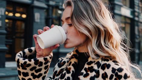 trendy blonde woman sipping takeaway coffee while walking along city sidewalk, wearing stylish leopard coat during sunny day with urban landscape blurring behind her