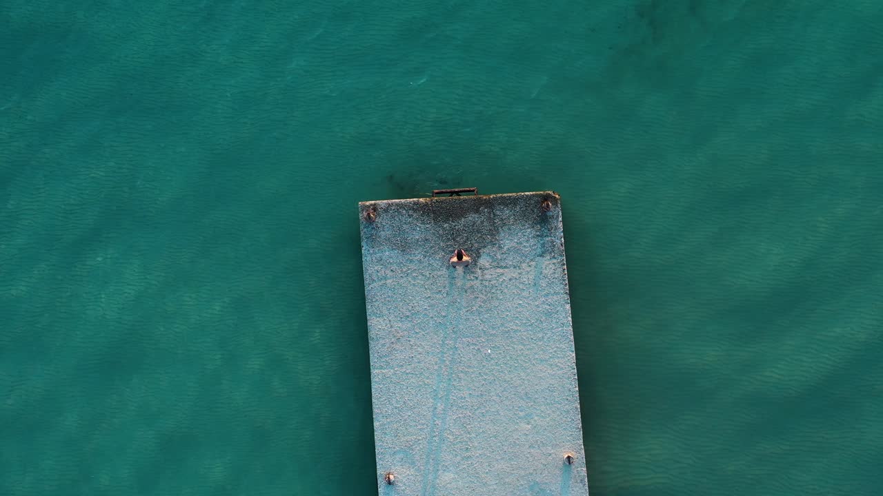 Ripresa con drone dall'alto di un uomo su un molo che guarda il mare blu cristallino.