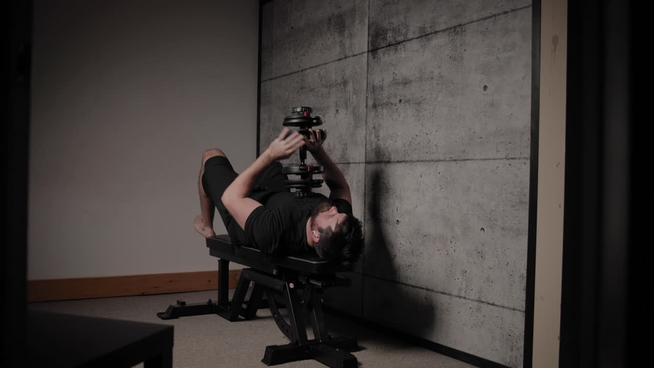 거짓말하는 덤벨 트라이세프 연장, 영화적인 조명, 검은색 체육복을 입은 백인 남자