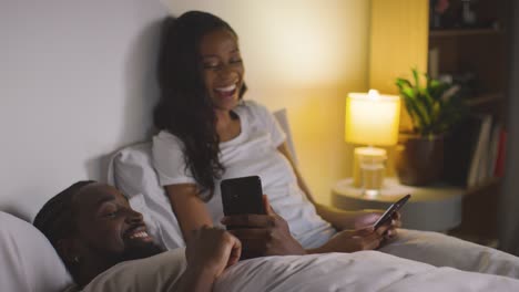 Smiling-Couple-At-Home-At-Night-Both-Looking-At-Mobile-Phones-In-Bed