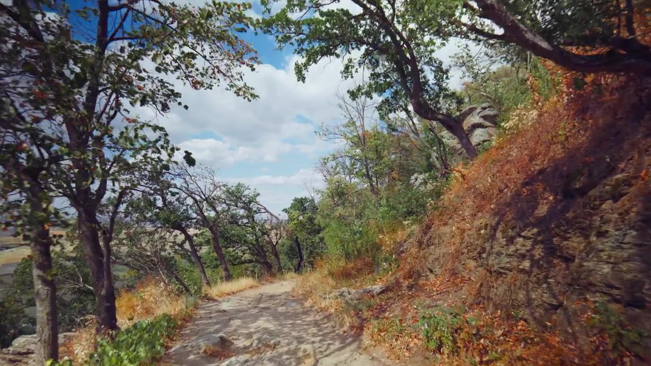 Ripresa video orizzontale con movimento fluido in avanti su un sentiero di montagna roccioso lungo una scogliera, con cielo blu vibrante e nuvole, paesaggi soleggiati e alberi naturali in una giornata luminosa