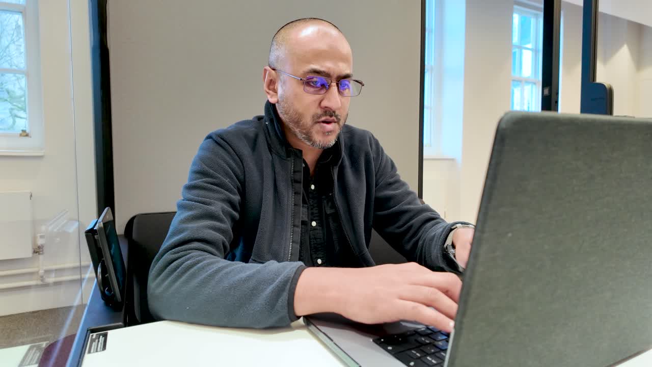 South Asian male professional typing on a laptop in a private office pod, showcasing focused work habits in a modern workspace