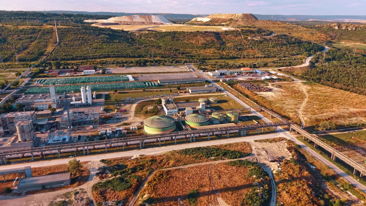 Industrial processing plant with pipelines below terraced quarry. High angle view of green vessels, pipe racks and service roads bordered by wooded hills.