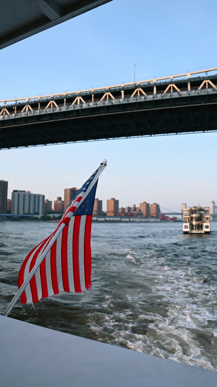 Barca con bandiera americana che si muove lungo il fiume. Angolazione dal basso verso il Manhattan Bridge con un treno in movimento. Video verticale