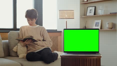 woman reading a book on sofa with laptop