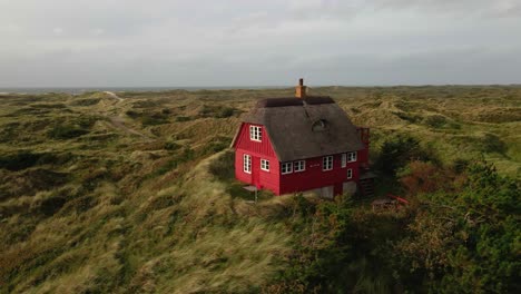 a drone orbits to the right around a red wooden house with a thatched roof at hvide sande beach, capturing the picturesque scene with stormy skies and sandy dunes in the background