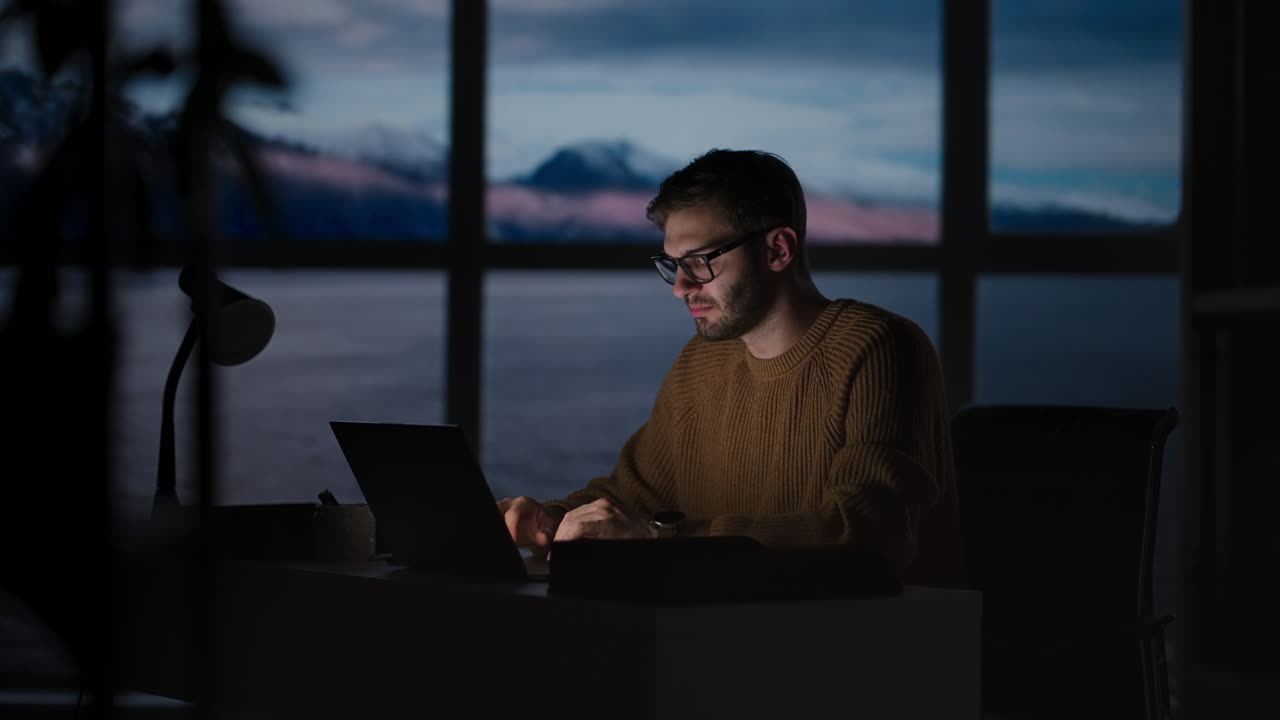 Tired young man working on a laptop late night in the office. Sleepy Businessman sitting at desk in dark office. Tired and stressed businessman in glasses works on a laptop of the night city office