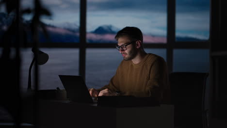 tired young man working on a laptop late night in the office. sleepy businessman sitting at desk in dark office. tired and stressed businessman in glasses works on a laptop of the night city office