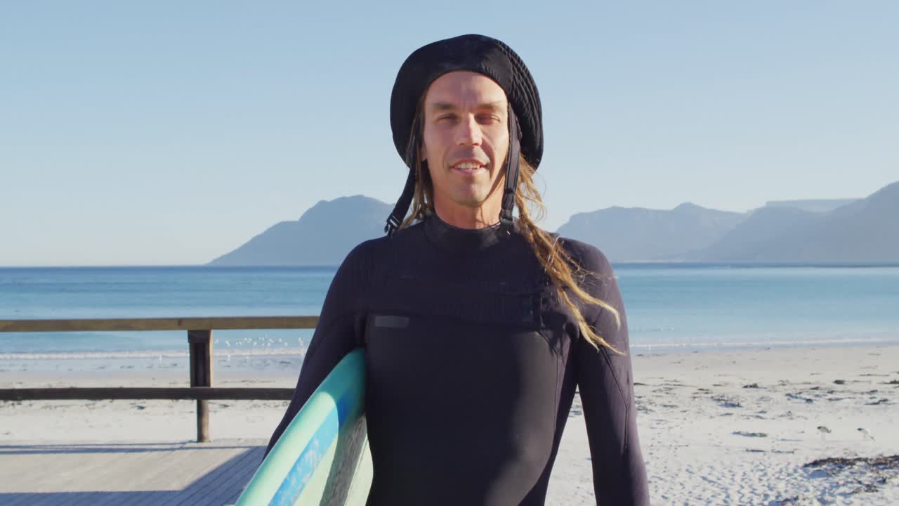 ⁇ 은 해변에서 서핑 보드를 들고 있는 웨이트 슈트에 드레드 로크를 입은 웃는 백인 남자의 비디오 초상화