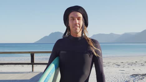 video portrait of smiling caucasian man with dreadlocks in wetsuit holding surfboard on sunny beach