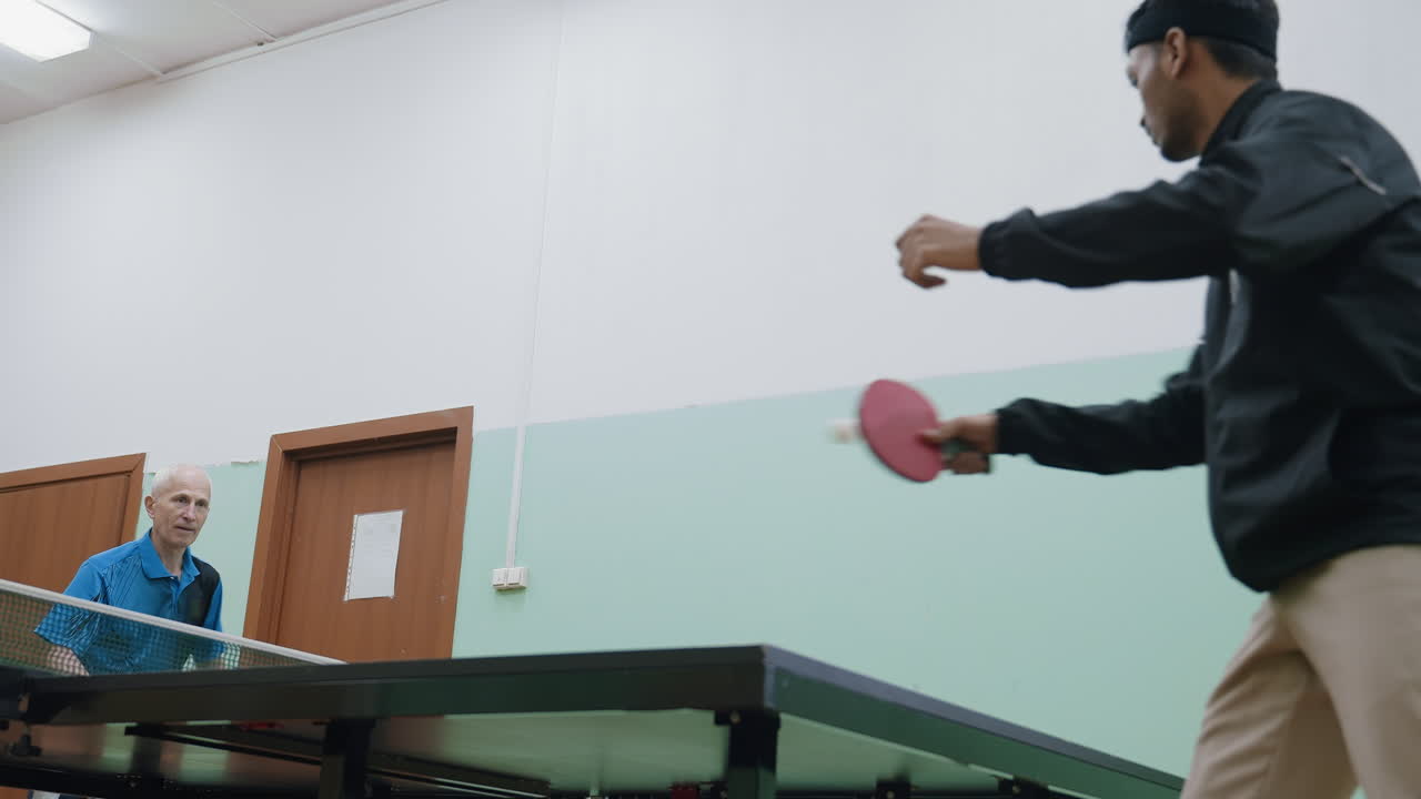 Seorang siswa muda dengan pakaian olahraga hitam berlatih tenis dengan pelatih senior dengan kemeja biru. Keduanya fokus pada reli di atas meja biru, bola putih bergerak di dalam ruang pelatihan dalam ruangan dengan pintu kayu