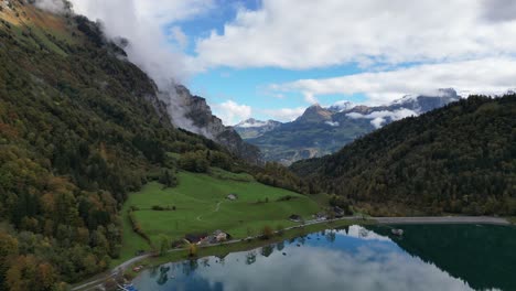 Aerial-shot-of-a-lake-and-grassland-within-the-valley-surrounded-by-high-mountain-peaks-and-trees