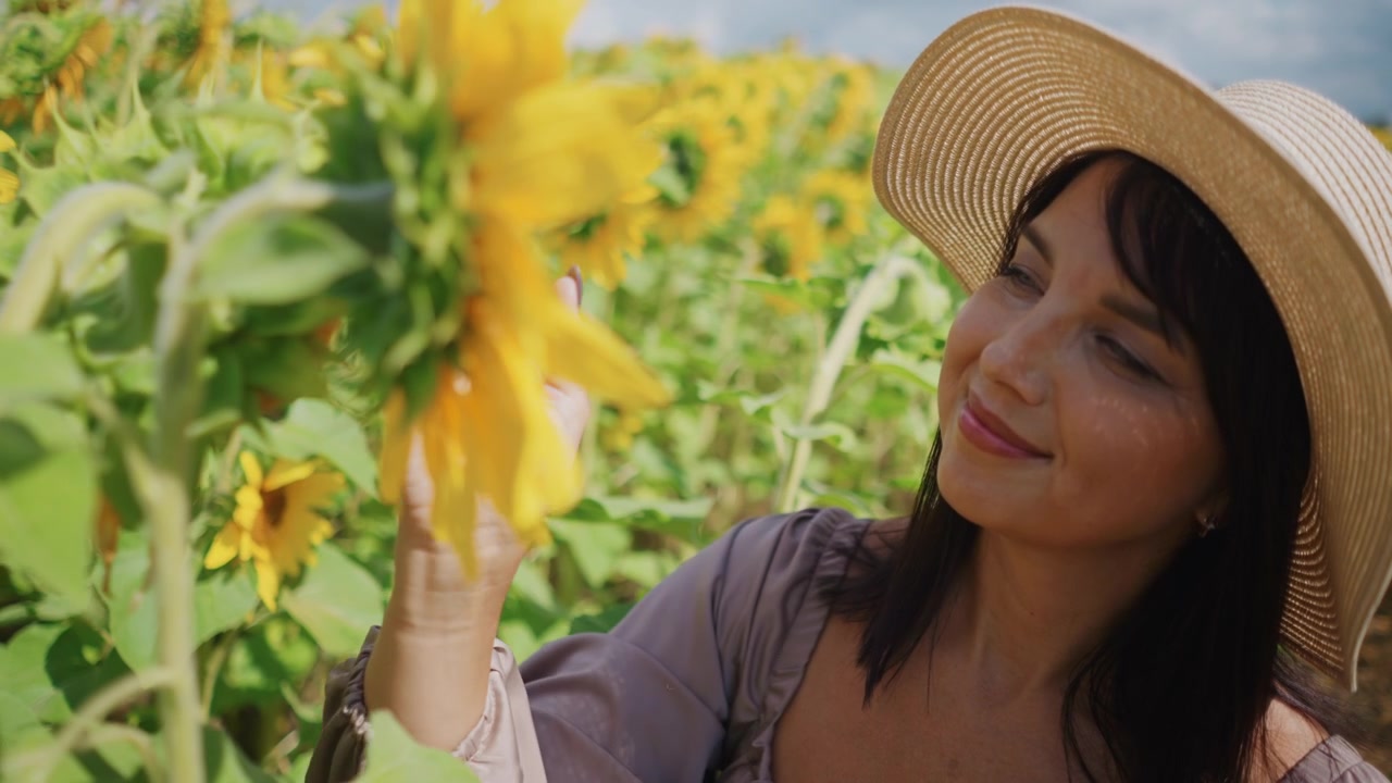 Primo piano di una donna con un cappello di paglia che tiene delicatamente ed esamina girasoli in piena fioritura. La scena presenta petali gialli vivaci, foglie verdi con texture visibile e illuminazione naturale soffusa.