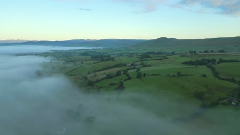 flying high over mist towards green patchwork fields of rural england at sunrise