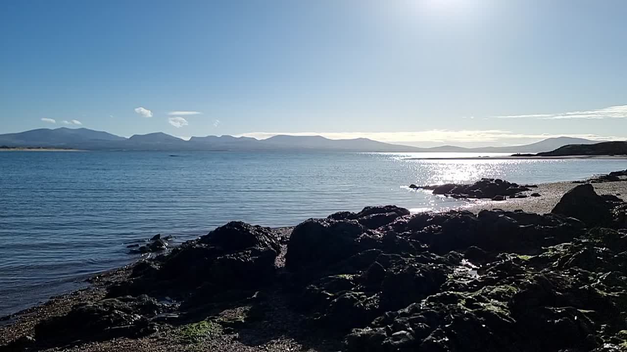 천천히 바위의  ⁇ 글세이 해안선을 가로지르고 해가 뜨면서 반 ⁇ 이는 바다 물과 스노우도니아 산맥의 지평선