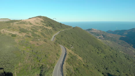 Aerial-shot-following-paraglider-along-mountain-road-to-distant-sea,-Liguria,-Italy,-Genoa