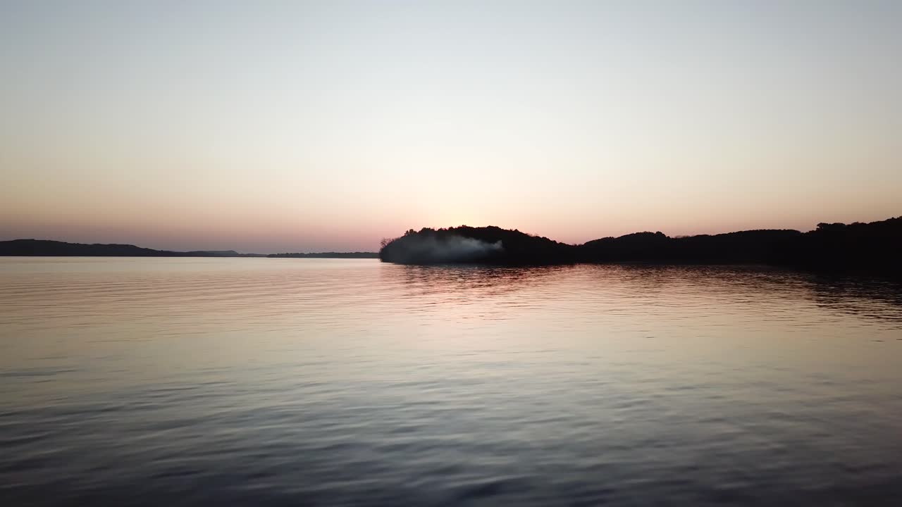 As dusk settles over Lake Wisconsin, a soft gradient sky reflects on the smooth water. Smoke drifts from a wooded peninsula, creating a tranquil and moody lakeside atmosphere.