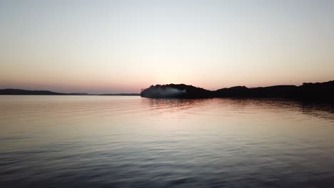 as dusk settles over lake wisconsin, a soft gradient sky reflects on the smooth water. smoke drifts from a wooded peninsula, creating a tranquil and moody lakeside atmosphere.