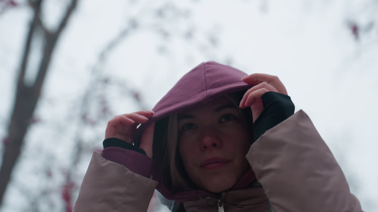 차가운 겨울 날씨에 은 코와 집중된 표정으로 머리 위에 후디를 당긴 젊은 여성, 따뜻함을 위해 후드를 조정하는 동안, 벌거벗은 나무 가지와 함께 흐릿한 얼어붙은 배경
