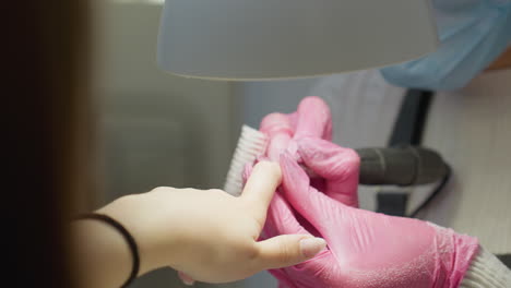 nail technician wearing pink gloves using electric filer and brush to clean and shape customer's fingernails during manicure session under bright salon lamp, with customer wearing black bracelet