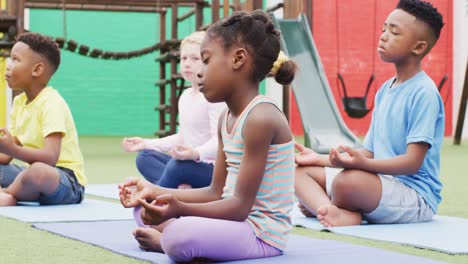 Diverse-schoolchildren-exercising-and-meditating-on-mats-at-school-playground