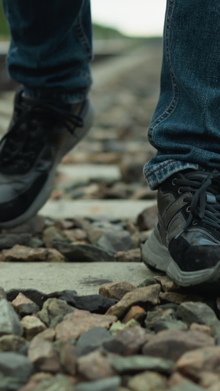 vista cercana de la pierna de alguien con pantalones vaqueros y zapatos de lona, caminando por un ferrocarril con piedras, el fondo incluye un borrón de árboles y postes eléctricos a ambos lados