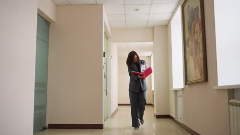 focused businesswoman walking through modern office hallway reviewing documents in red folder wearing dark formal suit with professional demeanor in bright workspace featuring framed painting
