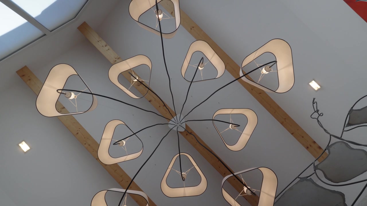 Lenta ripresa rotante con allontanamento di un moderno lampadario a più bracci con paralumi in tessuto triangolari arrotondati e cavi neri a vista che si irradiano dal supporto centrale su un soffitto bianco con travi in legno.