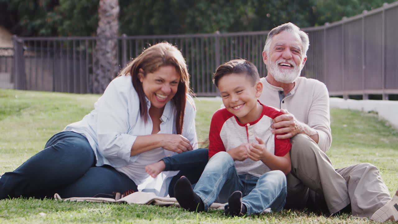Senior Hispanic couple sitting on grass tickling their grandchildren and smiling to camera