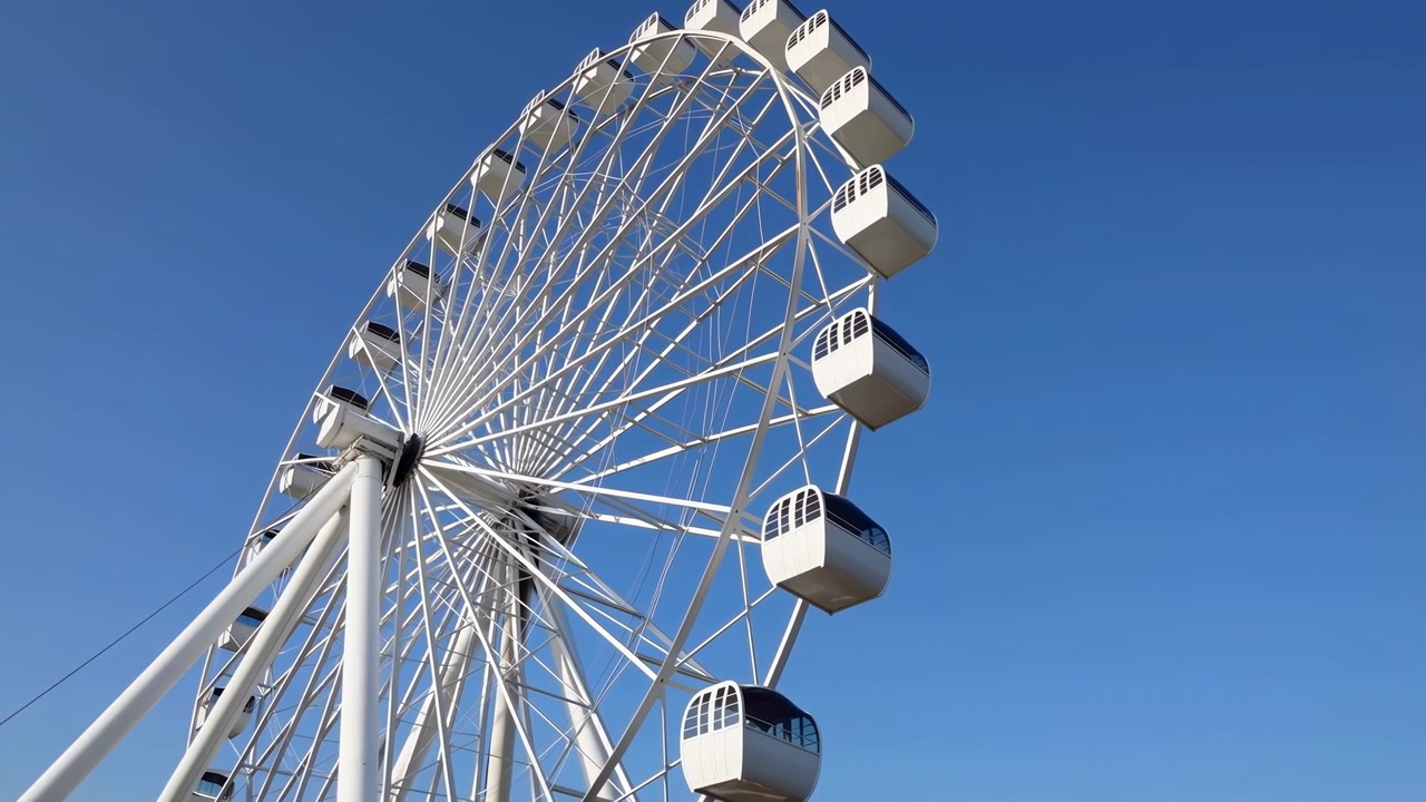 Ruota panoramica bianca contro cielo blu, inquadratura dal basso di gigantesca struttura in acciaio con cabine chiuse, raggi.