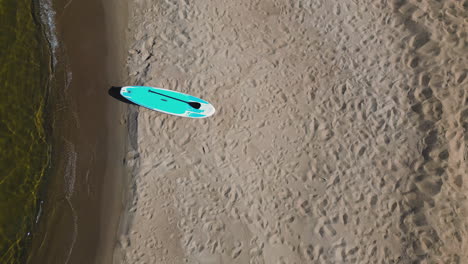 Aerial-view-of-lone-Stand-up-paddle-board-SUP-on-beach-at-the-edge-of-the-water