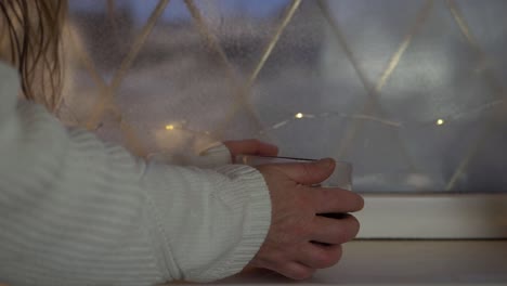 warming hands on a mug of hot coffee in winter window at home close up