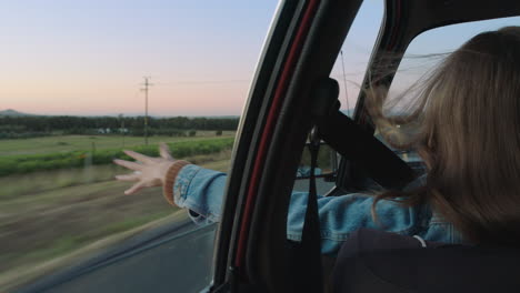 mujer joven en el coche con la mano afuera de la ventana sintiendo el viento soplando a través de los dedos conduciendo en el campo en un viaje por carretera viajando para las vacaciones de verano disfrutando de la libertad en la carretera al atardecer
