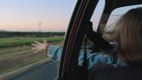 young woman in car holding hand out window feeling wind blowing through fingers driving in countryside on road trip travelling for summer vacation enjoying freedom on the road at sunset