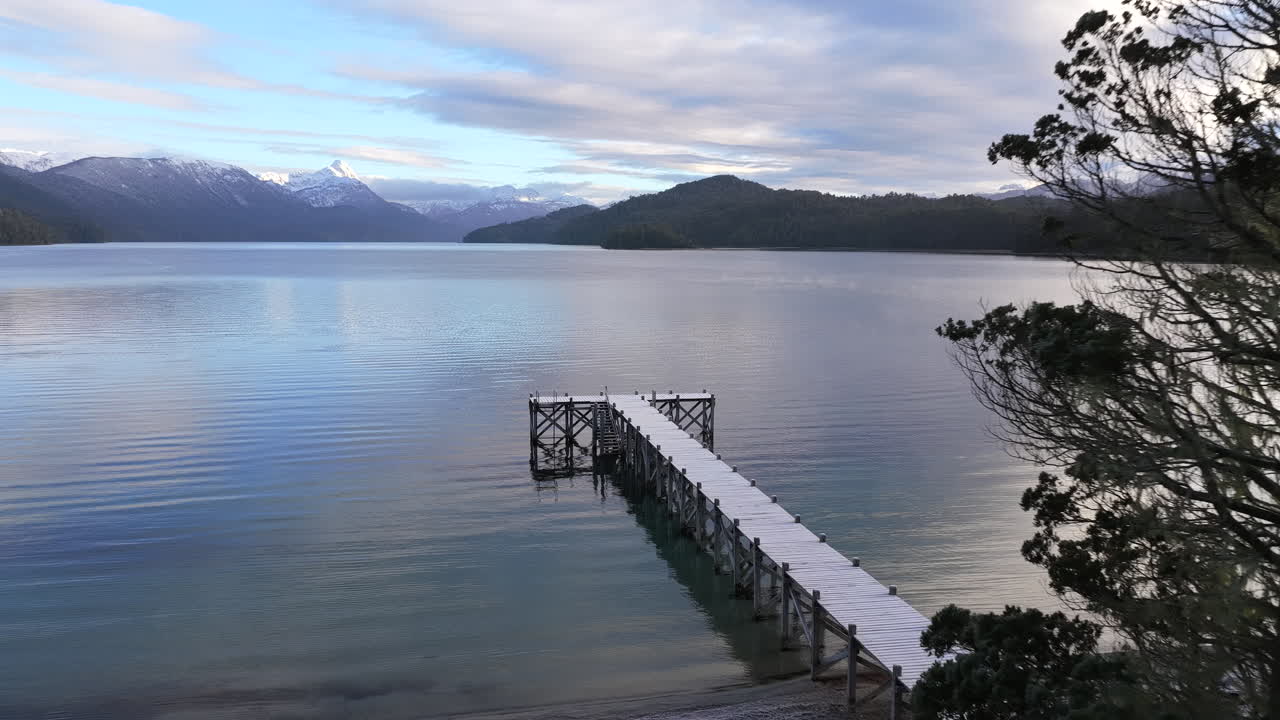 Un drone avanza verso un molo di legno sul Lago Espejo, con le montagne innevate delle Ande in Patagonia.