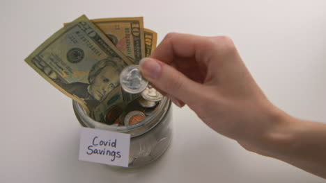 A caucasian woman&#039;s hand puts two American quarters into a &amp;quot;Covid Savings&amp;quot; jar