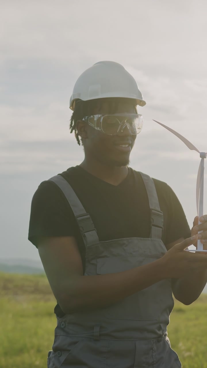 Man in hard hat and safety glasses examines wind turbine blades in a grassy field with a wind farm