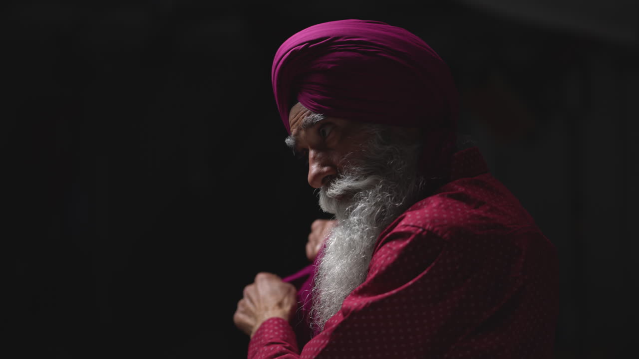 Close Up Low Key Studio Lighting Shot Of Senior Sikh Man With Beard Tying Fabric For Turban Against Dark Background Shot In Real Time