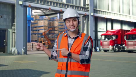 asian male engineer with safety helmet smiling and pointing to side while standing , outside of logistics distributions warehouse